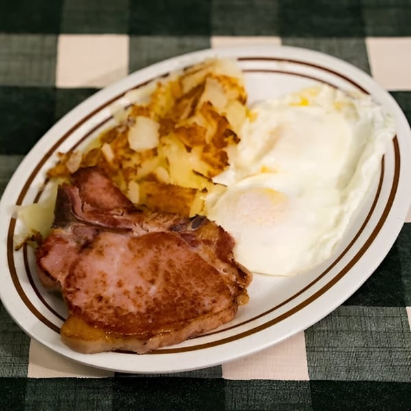 A diner breakfast of two fried eggs, hash browns, and a thick slice of ham on a white plate.