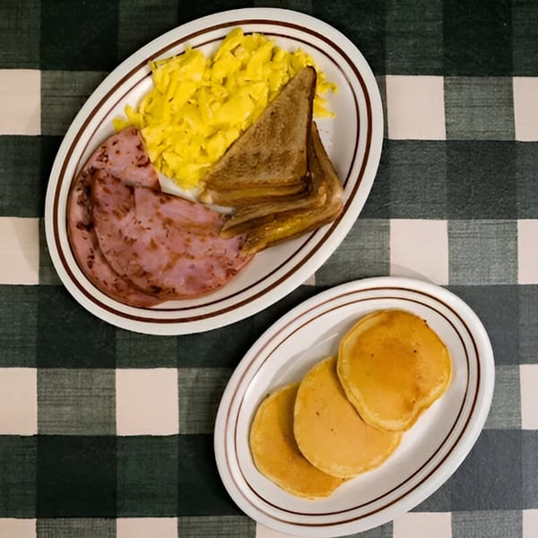Diner breakfast with scrambled eggs, ham, toast, and pancakes on a green and white checkered tablecloth.