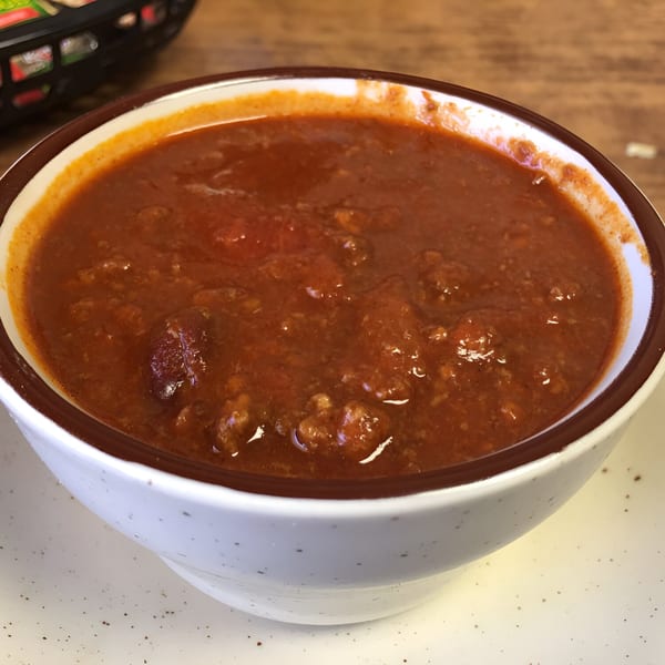A white bowl with a brown rim filled with Studebaker's hearty chili, on a speckled white plate.