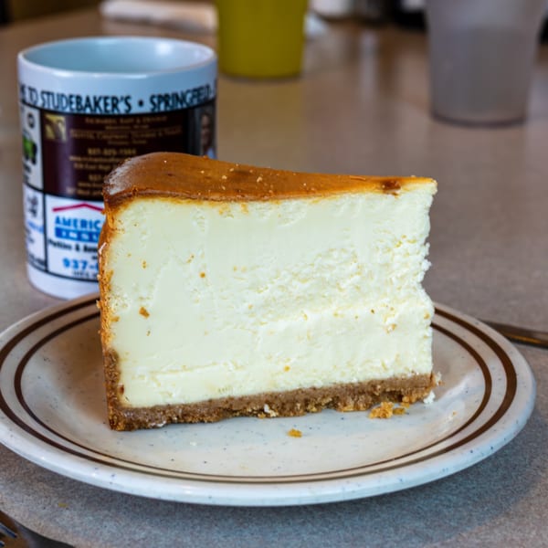A slice of cheesecake with a graham cracker crust on a diner plate next to a Studebaker's mug.