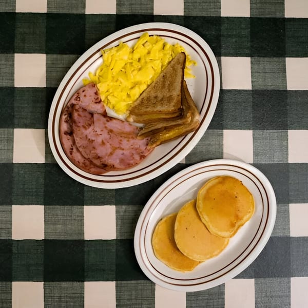 Diner breakfast with scrambled eggs, ham, toast, and pancakes on a green and white checkered tablecloth.